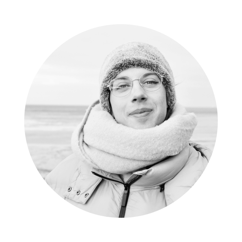 Ein schwarz-weiss Portrait von Ray. Sie steht am Meer und trägt Mütze, Schaal und eine dicke Winterjacke. Sie trägt eine Brille mit goldenem Rand und lächelt